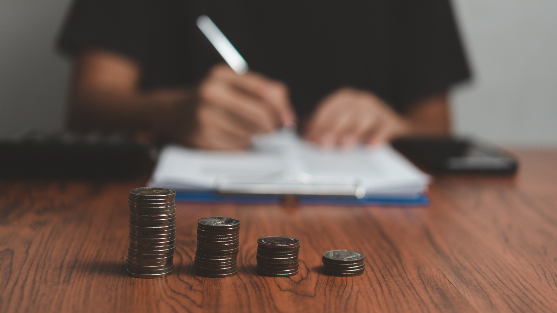Coin stacks of decreasing height on a wooden desk, a person filling in documents behind – E-commerce Manager salary