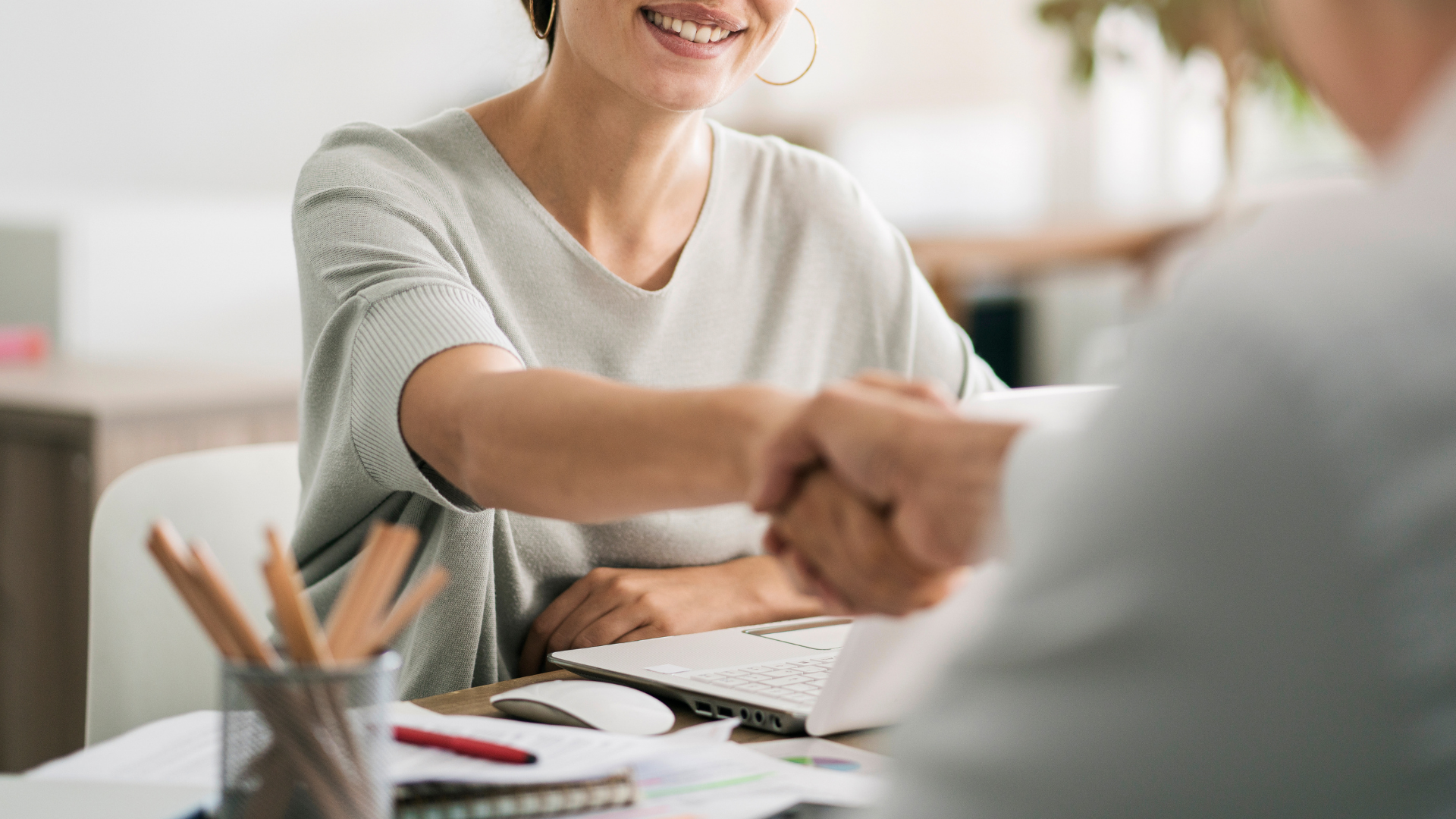 Smiling woman shaking hands with someone across a desk with a laptop – hiring an e-commerce specialist
